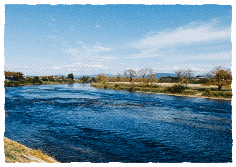 Una postal de un río ancho y azul entre orillas de hierba bajo un cielo pálido, con los bordes de la impresión rasgados